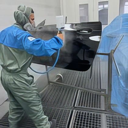 Ein Autolackierer in Schutzkleidung sprüht schwarzen Lack auf ein Fahrzeugteil.
