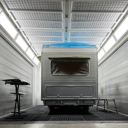 Wohnwagen in einer Lackierkabine während einer Hagelschadenreparatur, mit teilweise abgedecktem Dach und Arbeitsmaterialien auf einem Tisch daneben.